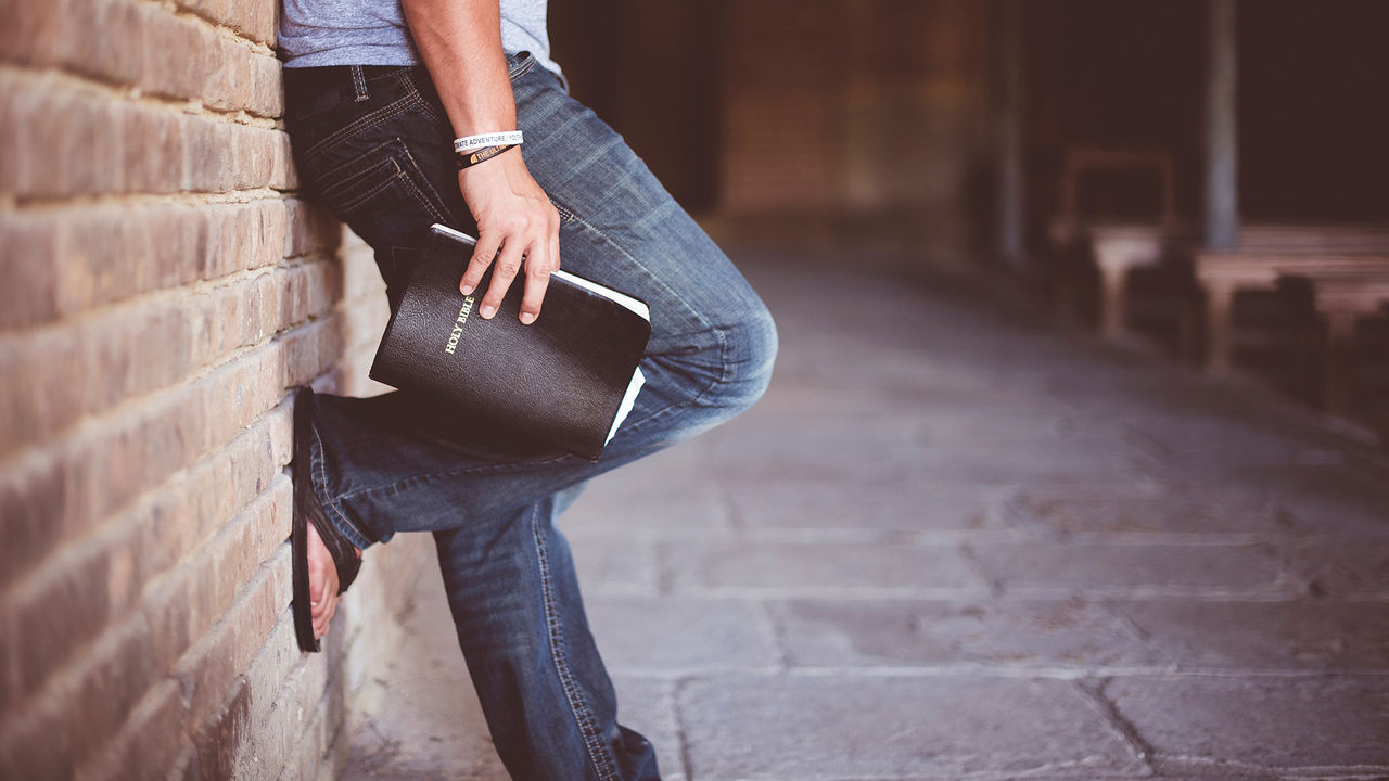 Man holding bible leaning against a brick wall.